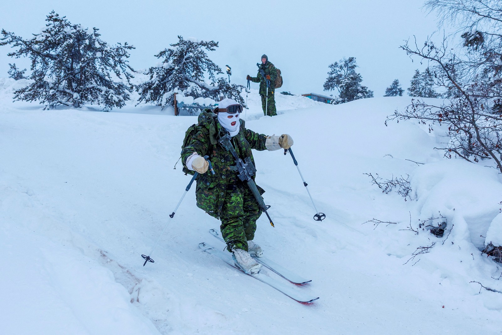 Soldiers wearing cold-weather gear and heavy packs ski across snowy ground.
