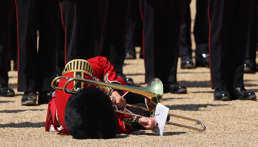 A soldier in dress uniform, holding a trombone, lies on the ground after falling.