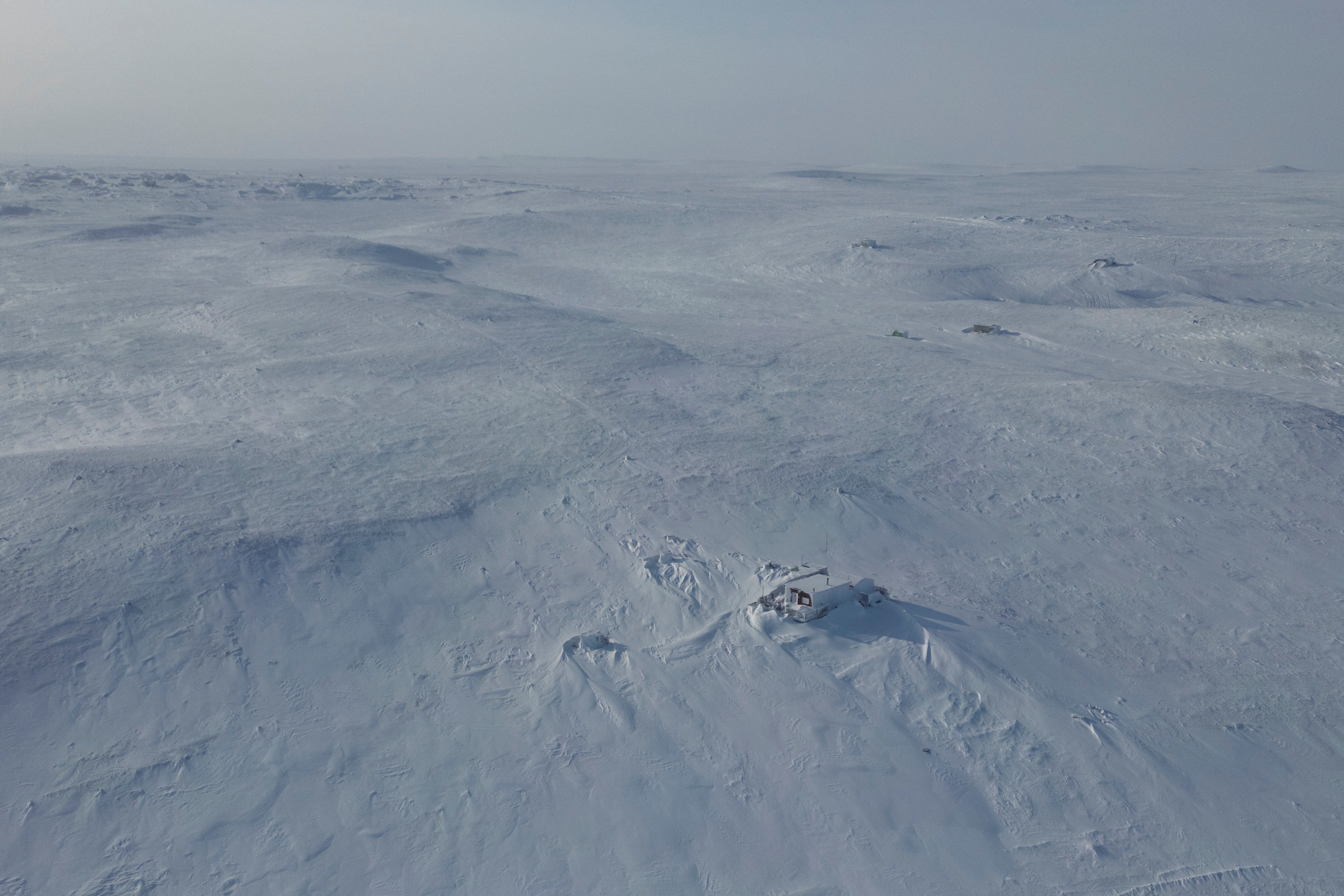 A drone view shows a structure partly buried in wind-blown snow in a vast plain of snow.