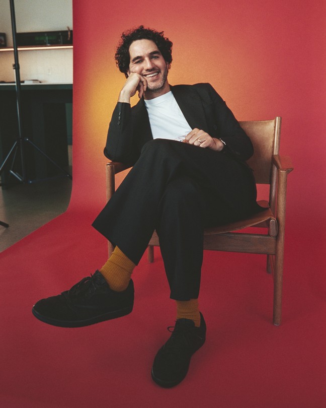 A color photograph of Benny Safdie seated in front of a red backdrop