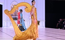A model walks on a stage wearing a costume shaped like a giant harp.