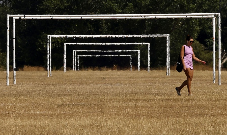 A person walks on dry grass past several sets of goalposts in a park.