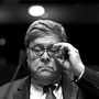 Shown in black and white, outgoing Attorney General Bill Barr adjusts his glasses while seated before a microphone