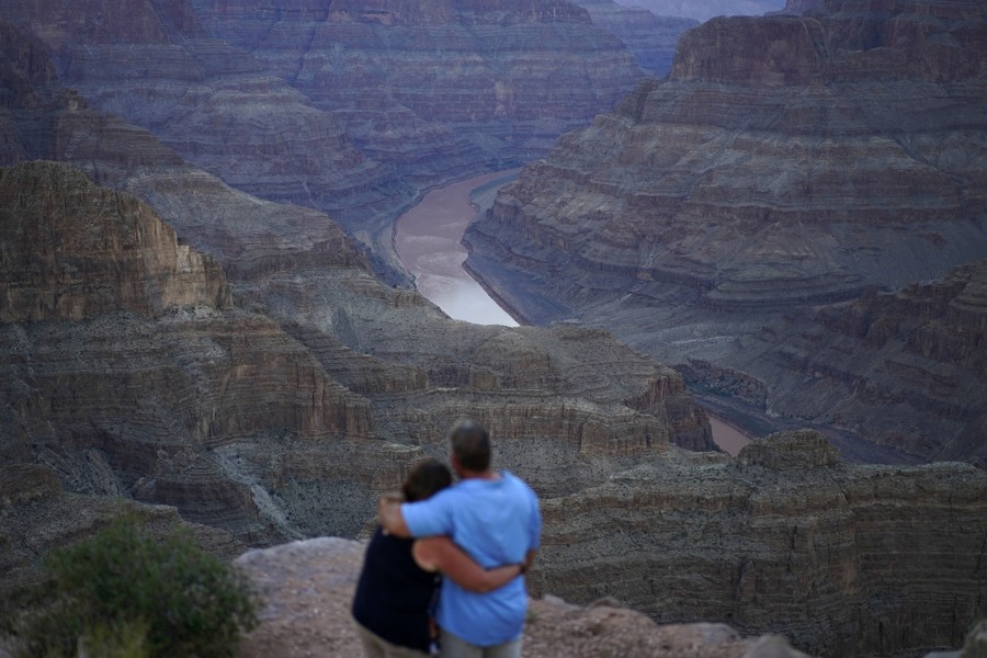 A couple embrace as they look over a valley with a river running through it.