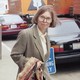 Color photo of Janet Malcolm, a white woman with brown bob in large round glasses and tan blazer, walking on city sidewalk with traffic in background.