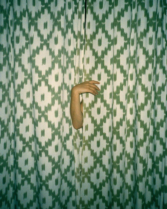Photograph of a curved arm reaching through green and white curtains