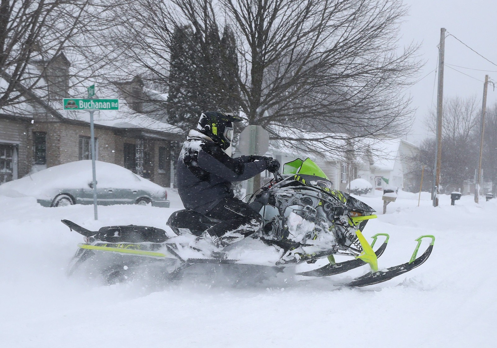 A person rides a snowmobile on a snow-covered road in a residential neighborhood.