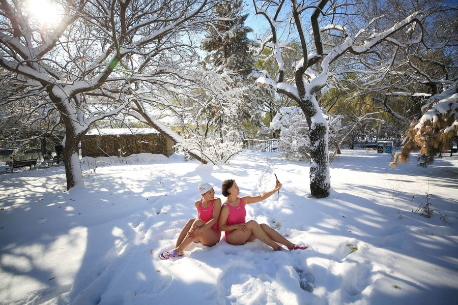 Two women in pink bathing suits sit in snow and take a selfie.