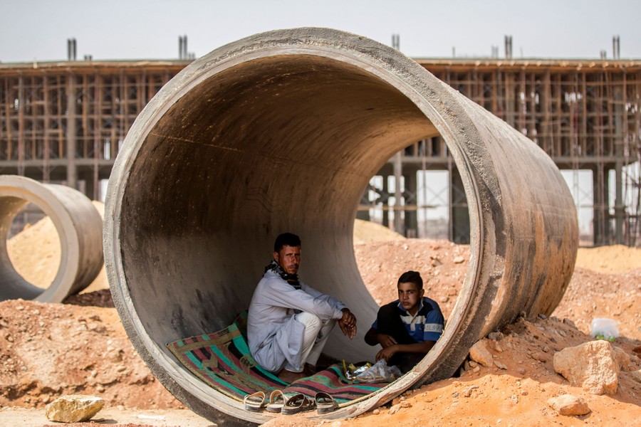 Two people sit and eat inside a large concrete pipe at a construction site.