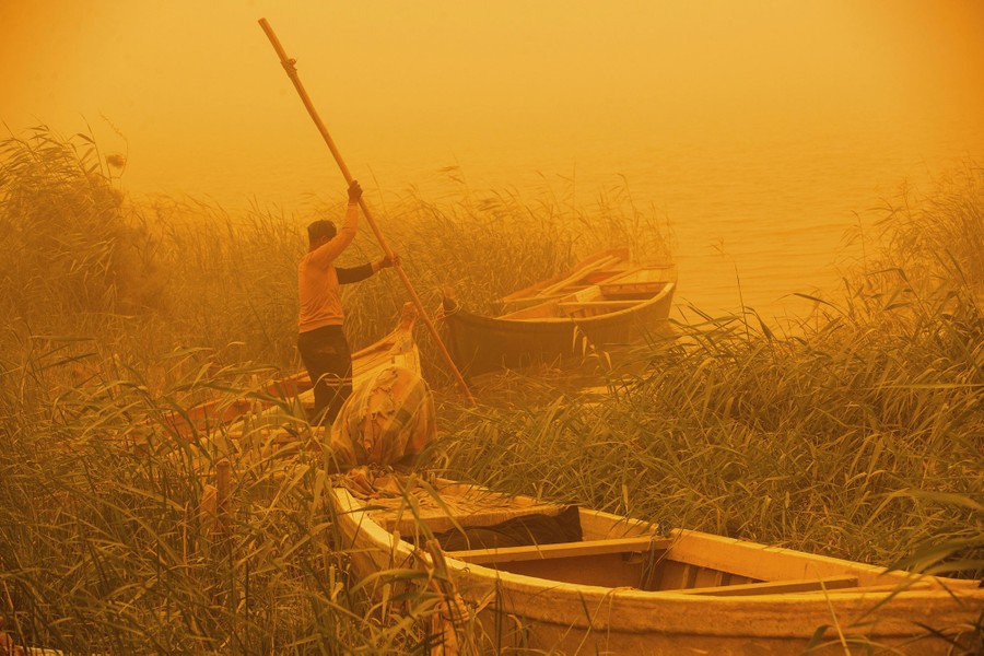 A person uses a pole to move a boat along a marshy river shoreline under an orange sky caused by a sandstorm.