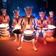 Five people in costume play illuminated drums in a street surrounded by a crowd.