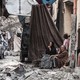 A family sits in the doorway of a ruined building, looking out on piles of rubble and a wrecked car.