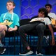 Several young spelling-bee contestants sit on chairs on a stage, with one appearing to be asleep.