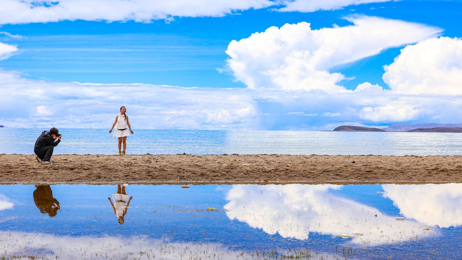 A person take a photo of another person beside a huge lake under a partly cloudy sky.