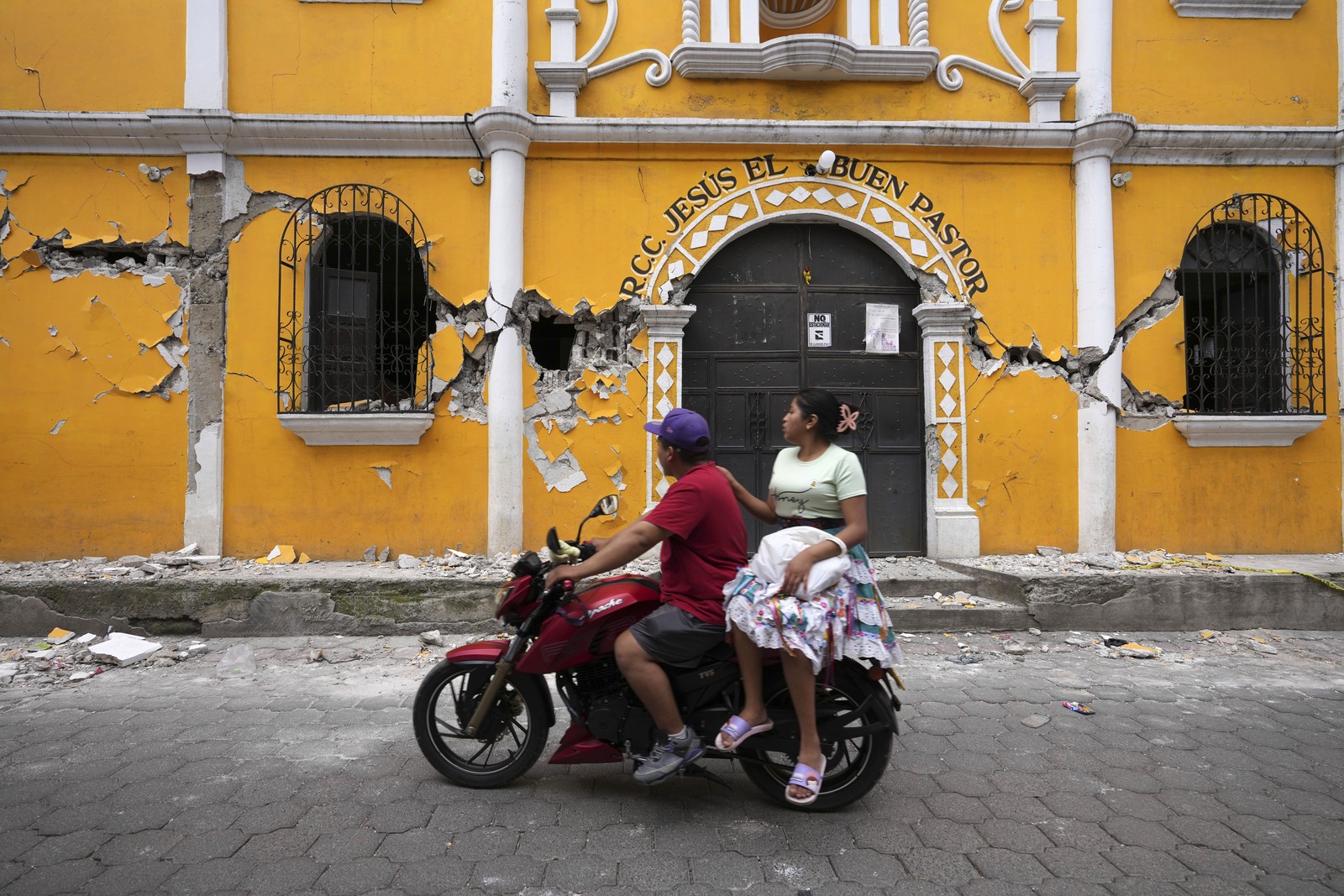 A couple rides a motorcycle past an older building that shows broad cracks after an earthquake.