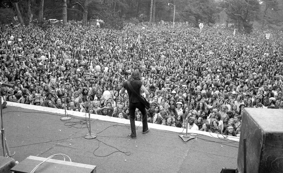 A large audience in a park watch a guitarist playing on a large stage.