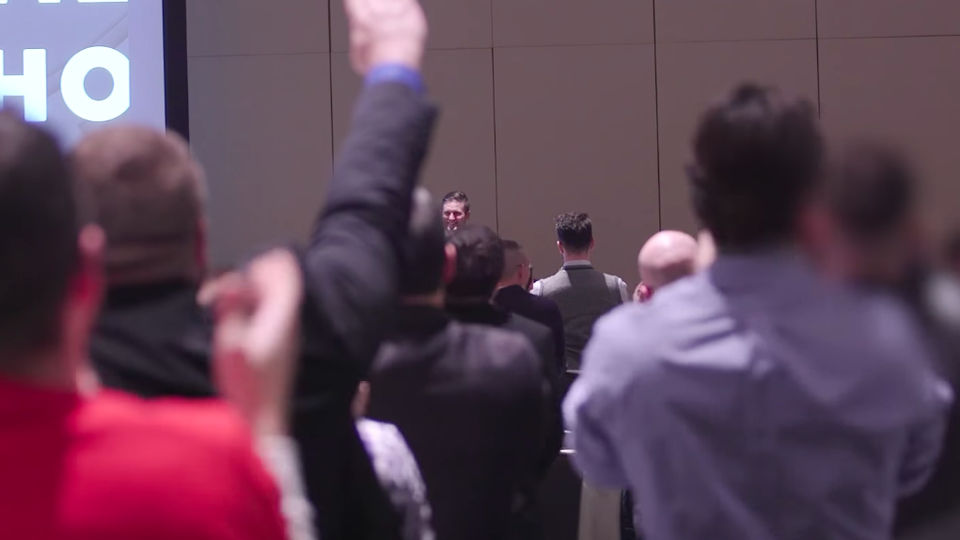 A crowd of white men seen from behind, one of whom has raised his arm in a Nazi-style salute