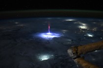 A nighttime view of the Earth, from orbit, with a bright blue-and-red bolt of electrical energy shooting up from a lightning strike in a storm below.
