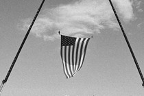 A black-and-white photograph of a large American flag being hung vertically over the stage of Donald Trump's Pennsylvania rally