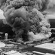 In a black-and-white photo, a large plume of smoke rises over buildings.