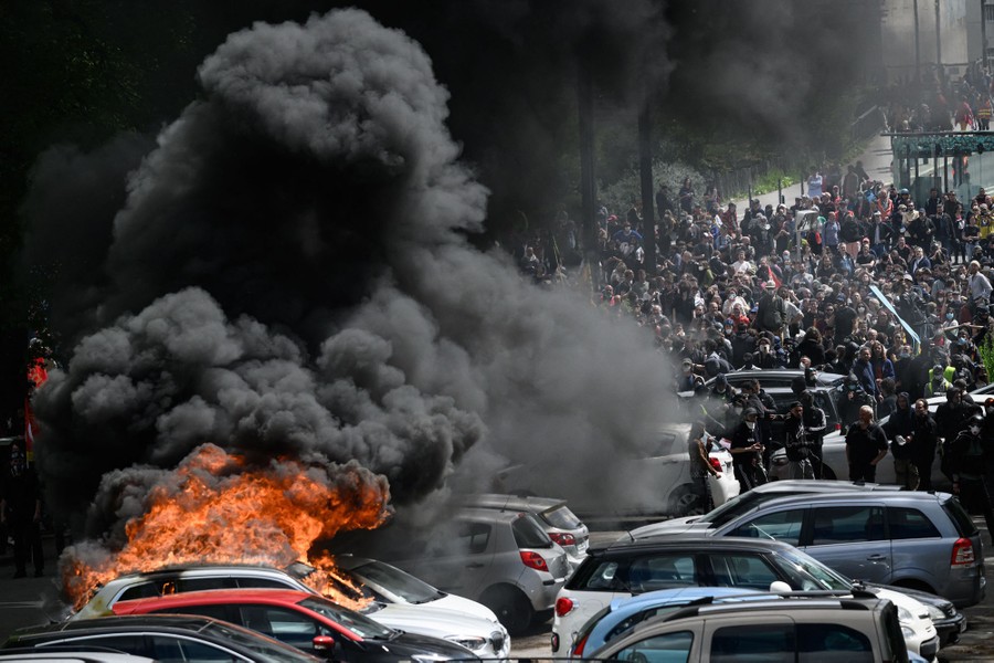 Several cars in a parking lot burn while protesters are gathered in the background.