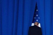 Color photograph of Donald Trump facing away from the camera, in front of the American flag and surrounded by a blue curtain.