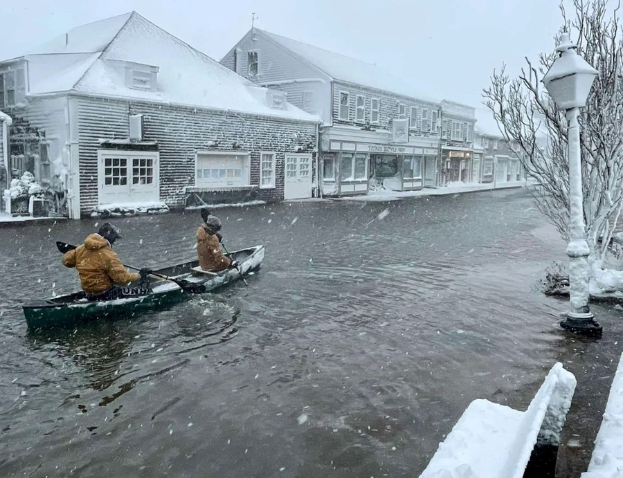 Two people row a canoe along a flooded road during a snowstorm.