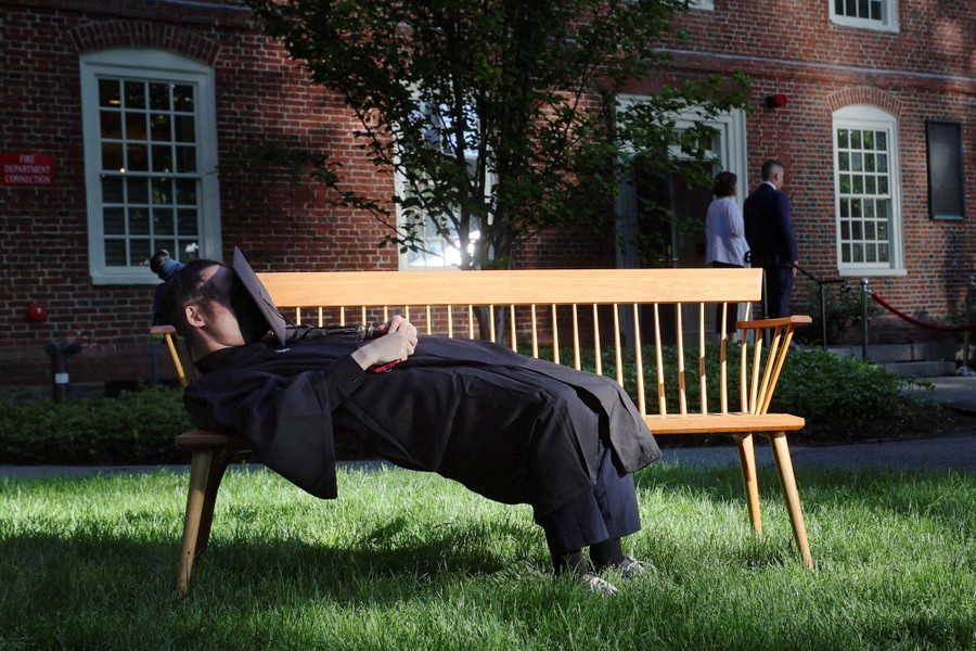 A student lies on a bench, wearing a gown and with a graduation cap placed over their face.