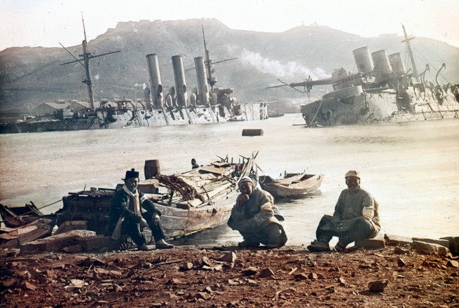 Three Japanese soldiers sitting on a beach near the wreckage of a partially sunken Russian warship.