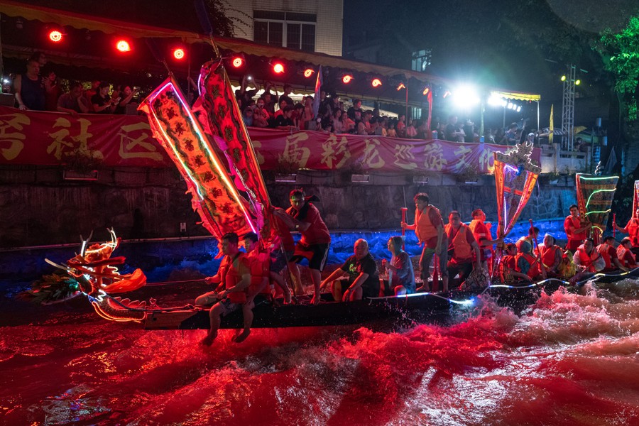 A crowd looks on while people ride and row in a long, thin boat decorated with a dragon's head on its bow.