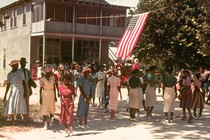 A photograph of group of Black people dressed in colorful summer celebration clothes in front of a hanging American flag and a two-story building