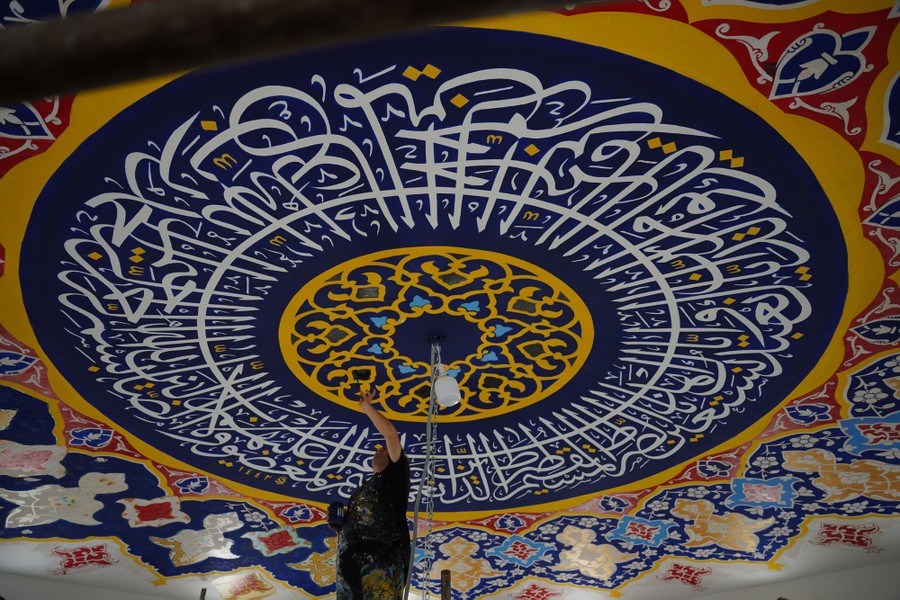 A person paints detailed Arabic writing onto the ceiling of a mosque.