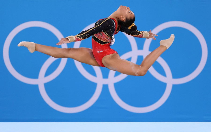 A gymnast leaps in front of a wall with the Olympic rings printed on it.