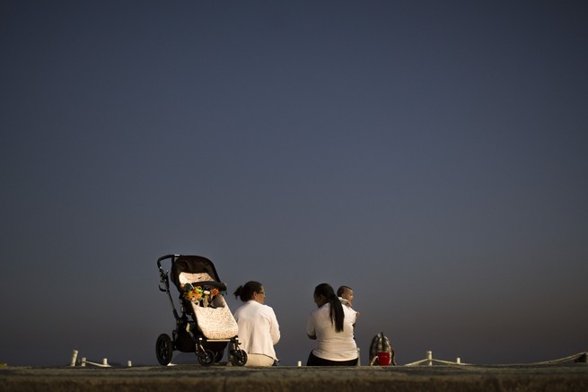 Two women sit on the ground, with their backs to the camera, looking off into the horizon. A black stroller is next to one woman, and the other is holding a child.