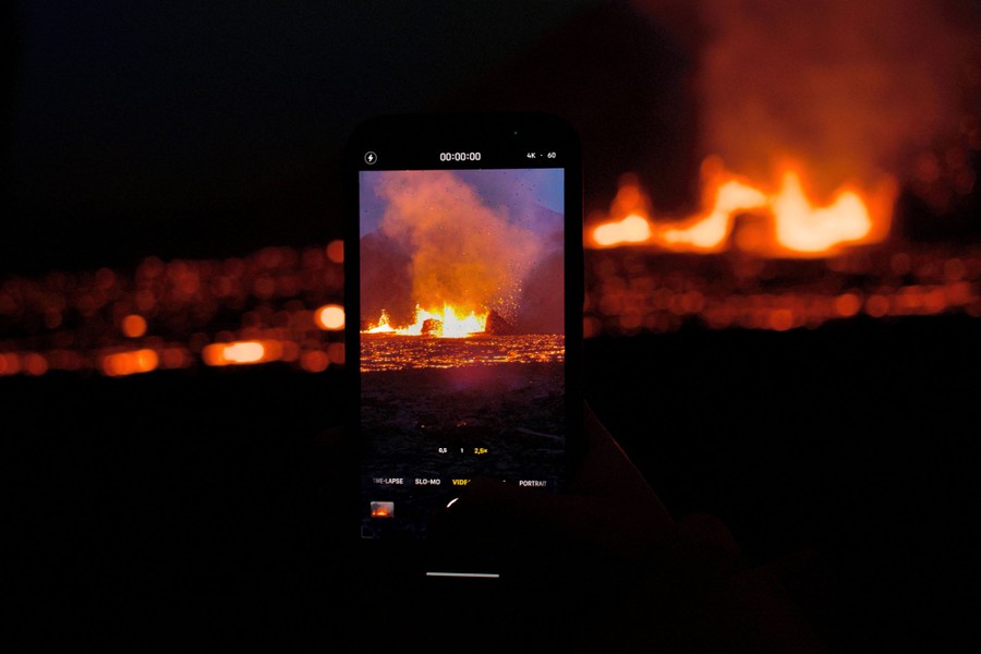 A person holds up a smartphone, taking a video of a volcanic-eruption scene behind it.