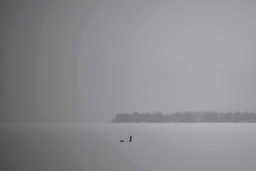 A distant view of a person pulling a sled on a snow-covered frozen body of water, under a gray-white sky.