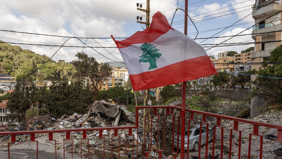 Photograph of a Lebanese flag waving on a red balcony overlooking the rubble of destroyed buildings across the street