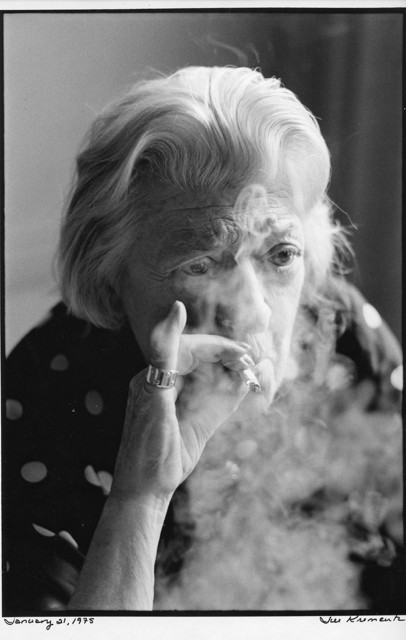 In a black and white photograph an elderly woman smokes a cigarette