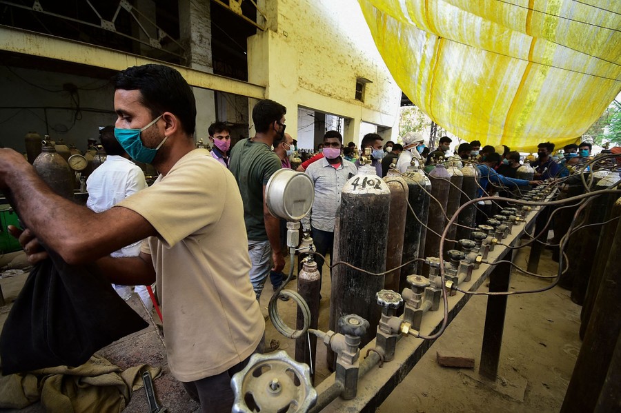 People gather and refill large tanks of oxygen near nozzles set up under a tarp.