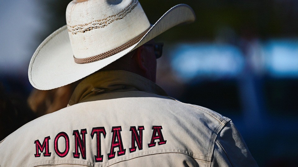 A photograph of a man from behind, wearing a white cowboy hat and a white jacket that says MONTANA in red font across the shoulders