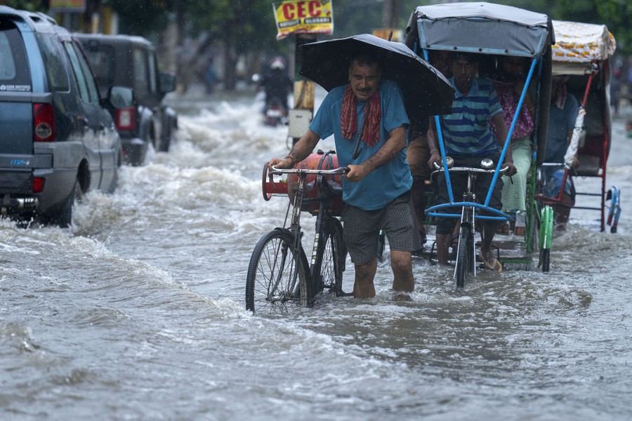 A man carries a gas cylinder on his bicycle while wading through a flooded street, among cars and rickshaws.