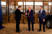 Elon Musk extends his hand to Donald Trump, who pats it. J. D. Vance stands next to them and smiles.