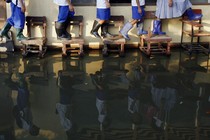 Students wearing rain boots walk on desks across their flooded classroom.