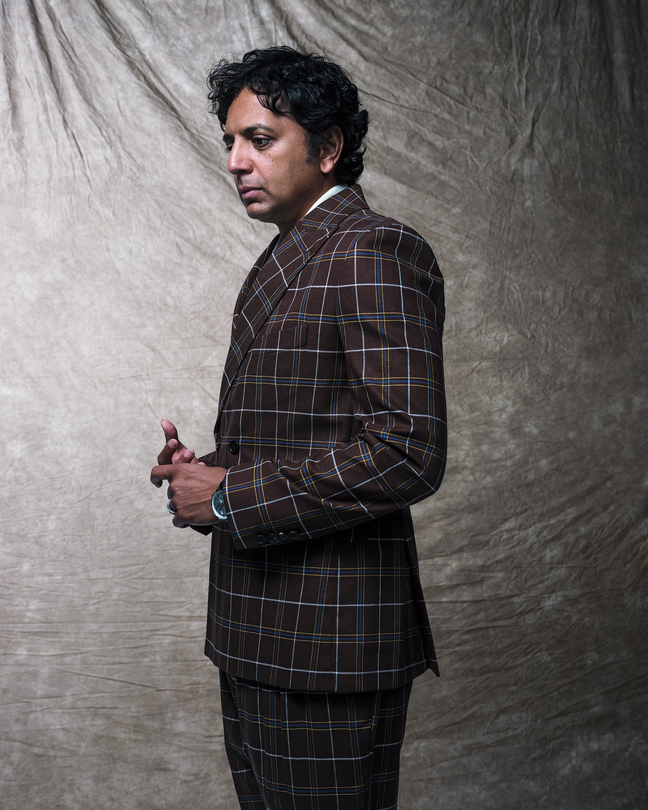 portrait of M. Night Shyamalan standing, looking down with hands clasped, wearing brown plaid suit