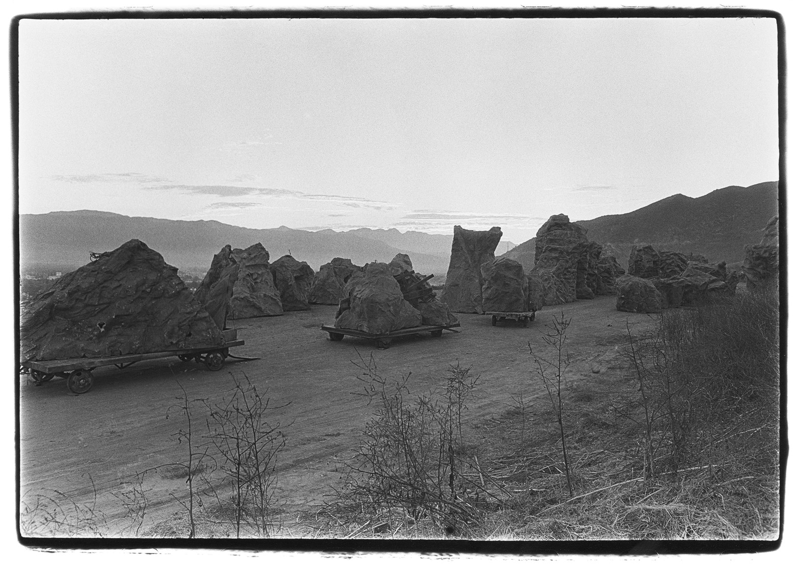 black-and-white photo of various large jagged boulders on moving dollies on dirt road with ridge of mountain in background
