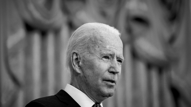 A black and white photo of Joe Biden. The background is blurry and the image cuts off at his shoulders. 