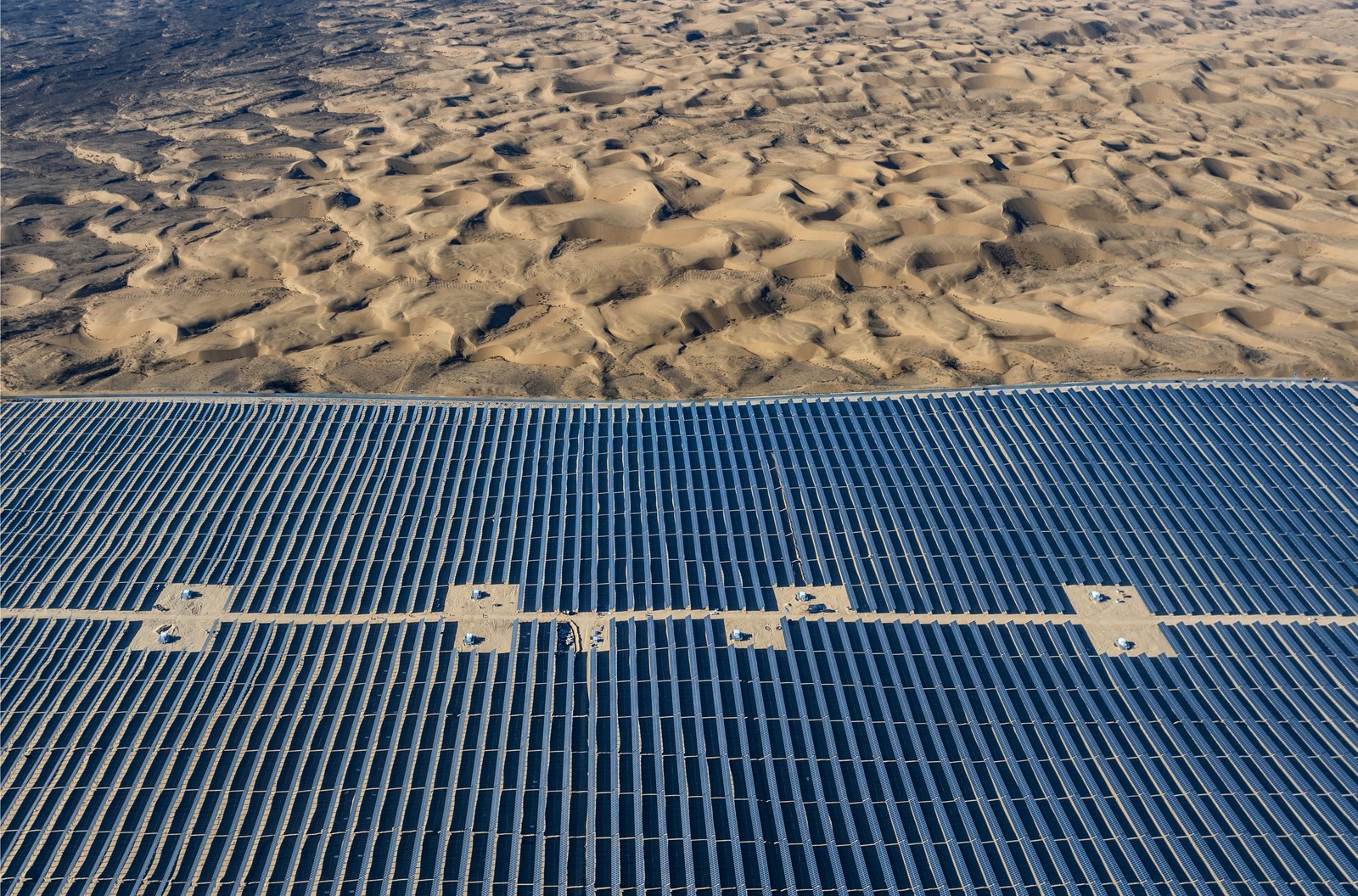 Many solar panels stretch across a desert, with dunes visible beyond.
