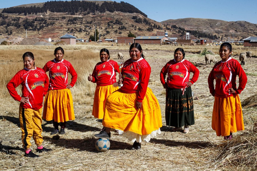 Indigenous women wearing red jerseys pose for a picture in a field. Many of them are wearing skirts; one has her foot on a soccer ball.