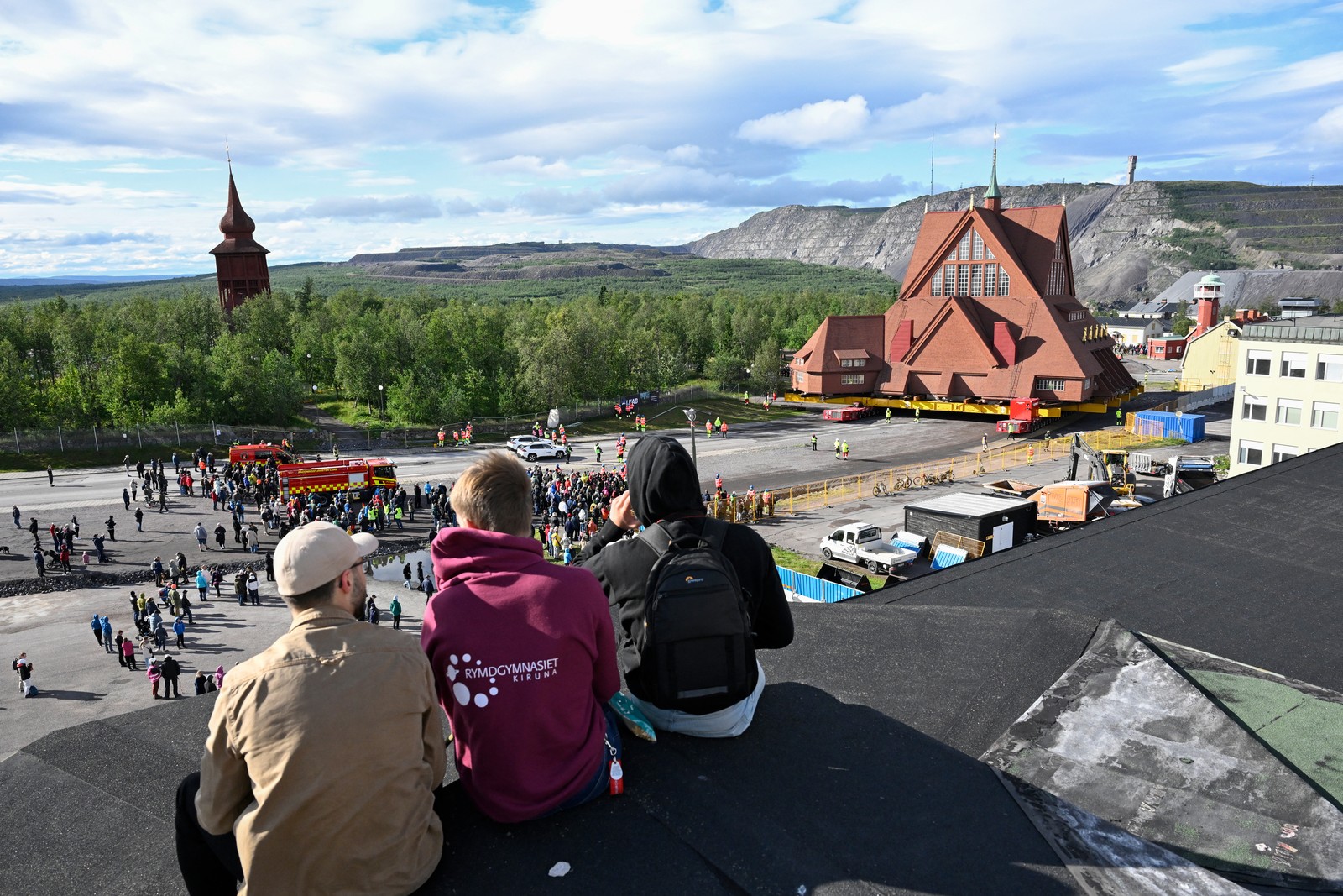 Several people sit on a rooftop, looking out toward a church being rolled slowly down a road.