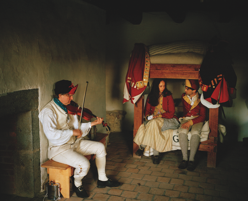 photo of room inside the English quarters of Fort Ticonderoga, where reenactors play a soldier playing the fiddle and two younger English residents sitting and talking on a wooden bed